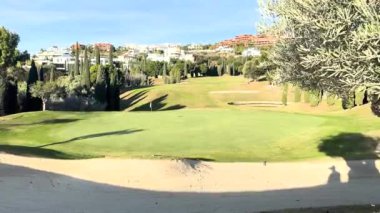 Arka planda yeşil kum sığınağı, ağaçlar ve lüks evleri olan manzaralı golf sahası güneşli mavi gökyüzünün altında..