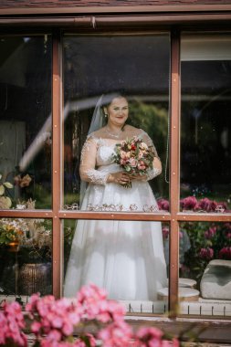 A bride in a lace wedding dress holds a bouquet while standing behind a window, framed by reflections, flowers, and greenery outside. clipart