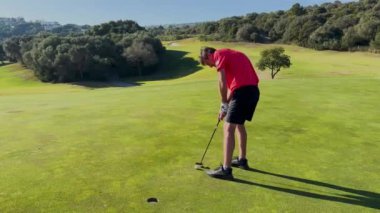 Sotogrande, İspanya - 26 Ocak 2024 - Biri yeşil üzerine kırmızı tişörtlü iki golfçü; diğeri manzaralı güneşli bir golf sahasının hizalama tavsiyesi için çömelirken diğeri bir vuruş hazırlıyor.