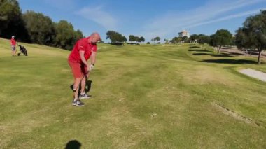 Sotogrande, İspanya - 26 Ocak 2024 - Kırmızı tişörtlü bir golfçü, arka planda ağaçlar, bir kum sığınağı ve bir kulüp binasıyla güneşli bir golf sahasına atış yapmaya hazırlanıyor..