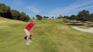 Sotogrande, İspanya - 26 Ocak 2024 - Kırmızı tişörtlü bir golfçü, arka planda ağaçlar, bir kum sığınağı ve bir kulüp binasıyla güneşli bir golf sahasına atış yapmaya hazırlanıyor..