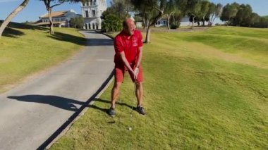 Sotogrande, İspanya - 26 Ocak 2024 - Kırmızı kıyafetli bir golfçü, arka planda ağaçlar ve kulüp binası olan güneşli bir golf sahasının yakınındaki bir araba yolunun yakınındaki çimlerden topu alır..