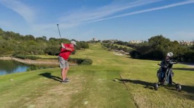 Sotogrande, İspanya - 26 Ocak 2024 - Kırmızı tişörtlü bir golfçü güneşli bir gökyüzünün altında ağaçlar, göletler ve uzak binalarla çevrili manzaralı bir golf sahasında sallanıyor.