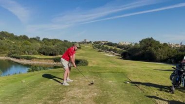 Sotogrande, İspanya - 26 Ocak 2024 - Kırmızı tişörtlü bir golfçü güneşli bir gökyüzünün altında ağaçlar, göletler ve uzak binalarla çevrili manzaralı bir golf sahasında sallanıyor.