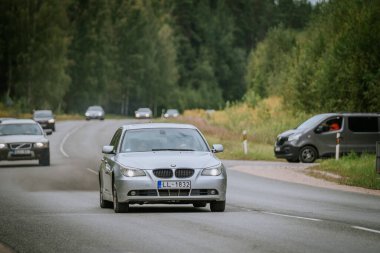 Valmiera, Letonya - 16 Ağustos 2024 - Arka planda diğer araçlar ve yol kenarına park edilmiş bir minibüsle birlikte orman yolunda giden gümüş renkli bir araba.