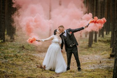 Valmiera, Letonya - 16 Ağustos 2024 - Gelin ve damat pembe duman bombaları tutarken romantik bir öpüşme paylaşıyorlar, canlı ve rüya gibi bir düğün sahnesi yaratıyorlar..