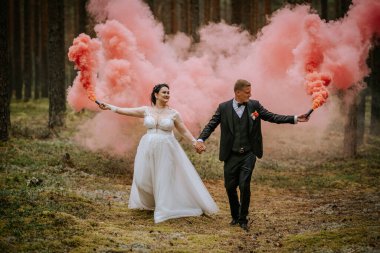 Valmiera, Letonya - 16 Ağustos 2024 - Gelin ve damat ormanda yürürken pembe duman bombaları tutarak eşsiz, romantik ve kaprisli bir düğün anı yarattılar.