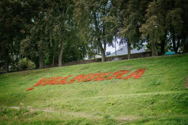 Valmiera, Latvia - August 16, 2024 - A green hillside features the word VALMIERA spelled out with vibrant red flowers, surrounded by trees and a serene outdoor environment clipart