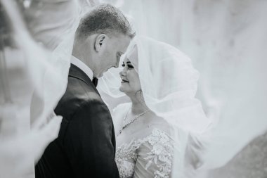 Valmiera, Letonya - 16 Ağustos 2024 - Bir gelin ve damadın romantik siyah beyaz portresi düğün günlerinde akan bir duvak altında hassas bir anı paylaşıyorlar.