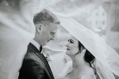 Valmiera, Letonya - 16 Ağustos 2024 - Bir gelin ve damadın akıcı bir peçenin altında romantik siyah beyaz portresi, düğün günlerinde birbirlerine sevgiyle bakıyorlar.