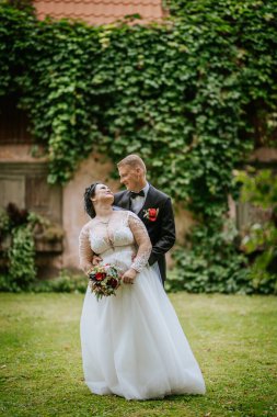 Valmiera, Letonya - 16 Ağustos 2024 - Beyaz elbiseli bir gelin ve damat açık havada, yeşil sarmaşık kaplı bir duvara karşı duran, elinde buket tutan romantik bir an paylaşıyorlar..