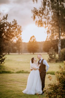 Valmiera, Letonya - 16 Ağustos 2024 - Bir gelin ve damat gün batımında açık havada, yemyeşil bir alanda ve arka planda romantik bir altın saat aydınlatmasıyla sevgi dolu bir anı paylaşıyorlar..