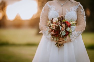 Dantelli gelinlik giymiş bir gelin, altın saat boyunca açık havada kırmızı, pembe ve yeşil çiçeklerden bir buket tutuyor ve romantik bir ortam yaratıyor..