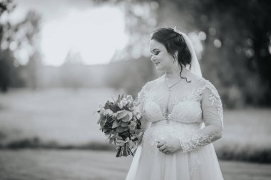 Elinde buket tutan bir gelinin siyah-beyaz portresi, zarif bir dantel elbise giymiş, açık havada duran, sakin bir arka planı olan.