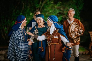 Valmiera, Latvia - August 16, 2024 - Women in traditional Latvian folk costumes talking and smiling, with a man playing a drum in the background during an outdoor cultural event. clipart