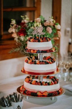 Valmiera, Latvia - August 16, 2024 - A three-tiered wedding cake decorated with fresh berries and a custom topper, displayed on a table with floral arrangements in the background. clipart