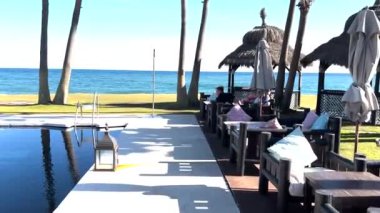 Sotogrande, İspanya - 25 Ocak 2024 - Güneşli bir günde okyanusa bakan sazdan şemsiyeli havuzu, palmiye ağaçları ve rahat oturma alanları olan tropikal sahil beldesi.