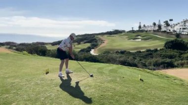 Sotogrande, İspanya - 26 Ocak 2024 - Bir golfçü, arka planda manikürlü çim yolları, sığınaklar ve yerleşim binaları bulunan denize bakan manzaralı bir pistte golf sopası sallıyor.