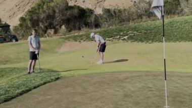 Sotogrande, İspanya - 26 Ocak 2024 - Yeşil üzerine iki golfçü, bir tanesi bayrağın yanına oturmaya hazırlanıyor, doğal yeşillik ve yakınındaki golf arabasıyla iyi korunan bir saha ile çevrili.