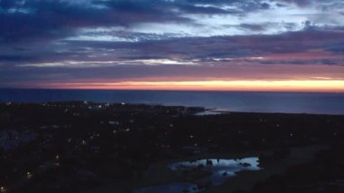 Alacakaranlıktaki kıyı golf sahasının havadan görünüşü su özellikleri, palmiye ağaçları, şehir ışıkları ve okyanusun üzerinde renkli bir günbatımı. İHA atışı.