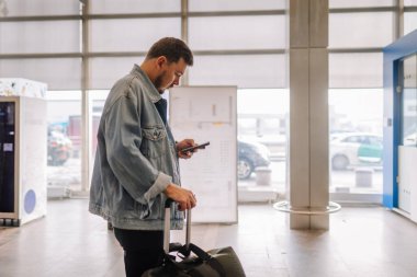 Riga, Letonya - 24 Ocak 2025 Havaalanında kot ceketli bir adam bagajı, arabaları ve arka planda görünen bir terminali olan telefonunu kontrol ediyor..