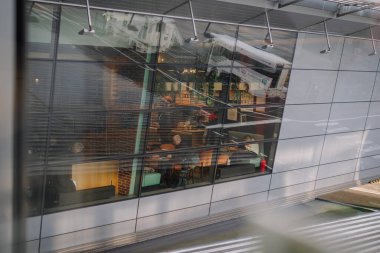 Riga, Letonya - 24 Ocak 2025 - Camdan bir binanın yansımaları, çevreci ve tuğla aksanıyla çevrili modern bir kafe içinde oturan insanların rahat bir atmosfer yarattığını gösteriyor..