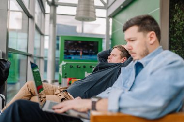 Riga, Latvia - January 24, 2025 -  Two men in a modern lounge, one focused on a laptop and the other reclining casually. Bright indoor space with green accents and a screen in the background. clipart