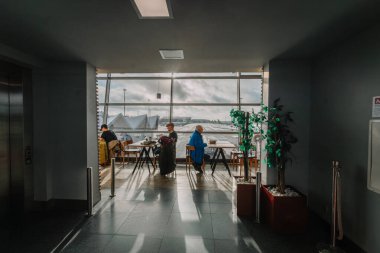 Riga, Latvia - January 24, 2025 - Airport seating area with people at tables, large windows with a view of the runway, potted plants, and natural light creating shadows. clipart