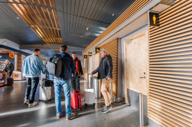 Riga, Latvia - January 24, 2025 - Travelers with luggage converse near restroom signs in a modern airport terminal featuring wooden panel walls and sleek design. clipart