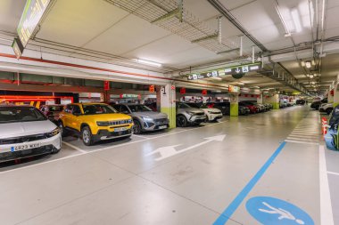 Malaga, Spain - January 24, 2025 - Underground parking lot with multiple cars, directional signs, and bright lighting, showing organized parking spaces and modern design. clipart