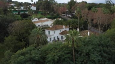  Kameralar yaklaştıkça yeşillik ve palmiye ağaçlarıyla çevrili havuzu, mobilyaları ve fayanslı çatısı olan lüks beyaz bir villanın insansız hava aracı videosu..