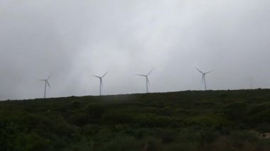 Rüzgâr türbinleri yeşil bir tepenin altında bulutlu, bulutlu bir gökyüzünün altında yenilenebilir enerji ve sürdürülebilirliği sembolize ediyor..