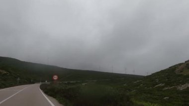Rüzgâr türbinleri arka planda bulutlu, bulutlu bir gökyüzünün altında yeşil tepelerin arasından geçen dolambaçlı bir yol yenilenebilir bir enerji manzarası yaratıyor..