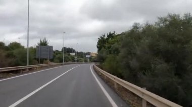 Etrafı yeşillik, bulutlu gökyüzü ve yol işaretleriyle çevrili, bulutlu bir günde sakin bir otoyol yolculuğunu tasvir eden kıvrımlı bir yol..