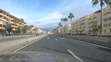 Sotogrande, İspanya - 26 Ocak 2025 - Güneşli bir gökyüzünün altında palmiye ağaçları ve bej apartman binalarıyla kaplı bir şehir caddesinde ilerlerken, trafik işaretleri görülebiliyor..