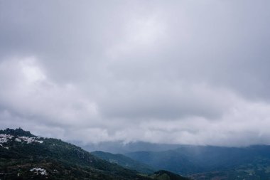 Gaucin, İspanya - 27 Ocak 2025 - Bulutlu bir gökyüzü, dağınık beyaz evler ve bereketli kırsal bir ortamda yemyeşil dağların dramatik manzarası.