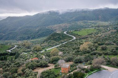 Gaucin, İspanya - 27 Ocak 2025 - Havadan dolambaçlı yollar, yeşil tepeler, dağınık ağaçlar, küçük kırsal bir bina ve bulutlu bir gökyüzü altında puslu dağlar.