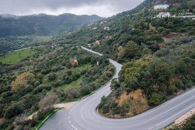 Gaucin, İspanya - 27 Ocak 2025 - Bereketli yeşil tepeler, yoğun bitki örtüsü ve bulutlu bir gökyüzü altında dağınık kırsal evlerle çevrili dolambaçlı bir yolun havadan görünüşü.