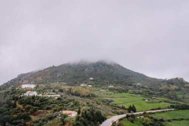 Gaucin, İspanya - 27 Ocak 2025 - Evleri, verimli yeşillikleri ve bulutlu bir gökyüzünün altında kısmen sisle kaplanmış bir dağ manzarası.