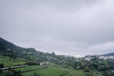 Gaucin, İspanya - 27 Ocak 2025 - Yeşil tepelerin, dağınık evlerin, dolambaçlı yolların ve bulutlu bir gökyüzünün altında sakin bir kırsal bölgenin havadan görünüşü.