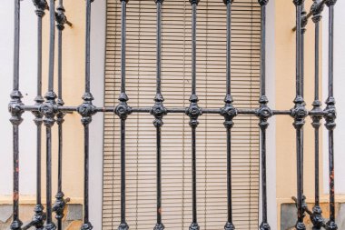 Gaucin, İspanya - 27 Ocak 2025 - İspanya 'nın Gaucin şehrinde bej bir duvar ve pencere perdelerine dayanan, detaylı detaylandırılmış siyah işlemeli demir pencere ızgarasına yakın plan.