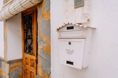 Gaucin, İspanya - 27 Ocak 2025 - İspanya Gaucin, İspanya 'da işlenmiş demirle döşenmiş ahşap bir kapının yanındaki duvarda dekoratif bir logosu olan beyaz bir posta kutusunun yakın çekimi.