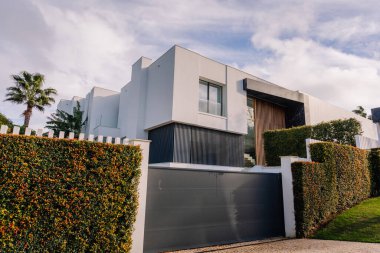 Sotogrande, Spain - January 27, 2025 - Contemporary white house with large windows, wooden accents, a dark gate, lush hedges, and a palm tree under a sunny blue sky. clipart