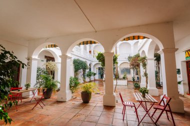 Sotogrande, İspanya - 27 Ocak 2025 - Beyaz kemerleri, terakotta fayansları, saksı bitkileri ve ahşap bir masanın etrafına kırmızı sandalyeler serin bir Akdeniz atmosferi yaratan zarif bir avlu.