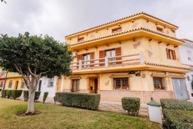 Sotogrande, İspanya - 27 Ocak 2025 - Ahşap panjurları, balkonları, fayanslı cephesi, çalıları, bir ağacı ve banliyö bölgesinde bulutlu bir gökyüzünün altında küçük bir çimenliği olan kırsal sarı bir ev.