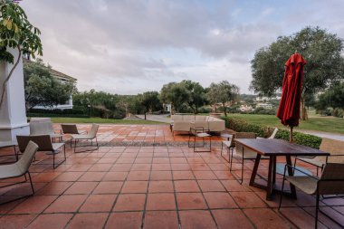 Sotogrande, İspanya - 27 Ocak 2025 - Terasotta fayansları, modern mobilyalar, kırmızı şemsiye ve yeşil, ağaçlar ve uzak binaların manzaralı terası.