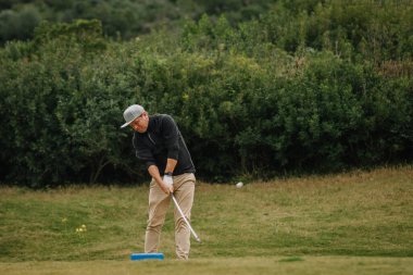 Yeşilliklerle çevrili çimenli bir golf sahasında sallanan bir golfçü, golf topuna hassasiyetle vurmaya odaklanıyor..