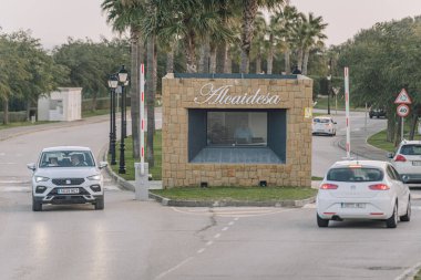 Sotogrande, Spain - January 29, 2025 - A gated entrance to Alcaidesa with a stone guardhouse, cars passing through, and palm trees lining the road in a tranquil setting. clipart