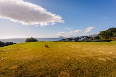 Endülüs, İspanya, 30 Ocak 2025 - Açık mavi gökyüzü, yamalı yeşil ve manzaralı okyanus manzaralı, yeşil ve beyaz binalarla çevrili kıyı golf sahası.