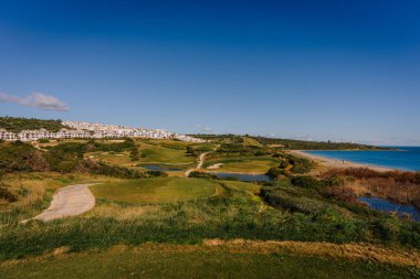 Endülüs, İspanya, 30 Ocak 2025 - La Hacienda golf sahasının panoramik manzarası, yemyeşil alanlar, su hatları, modern konutlar ve açık mavi gökyüzü altında kumlu bir sahil.